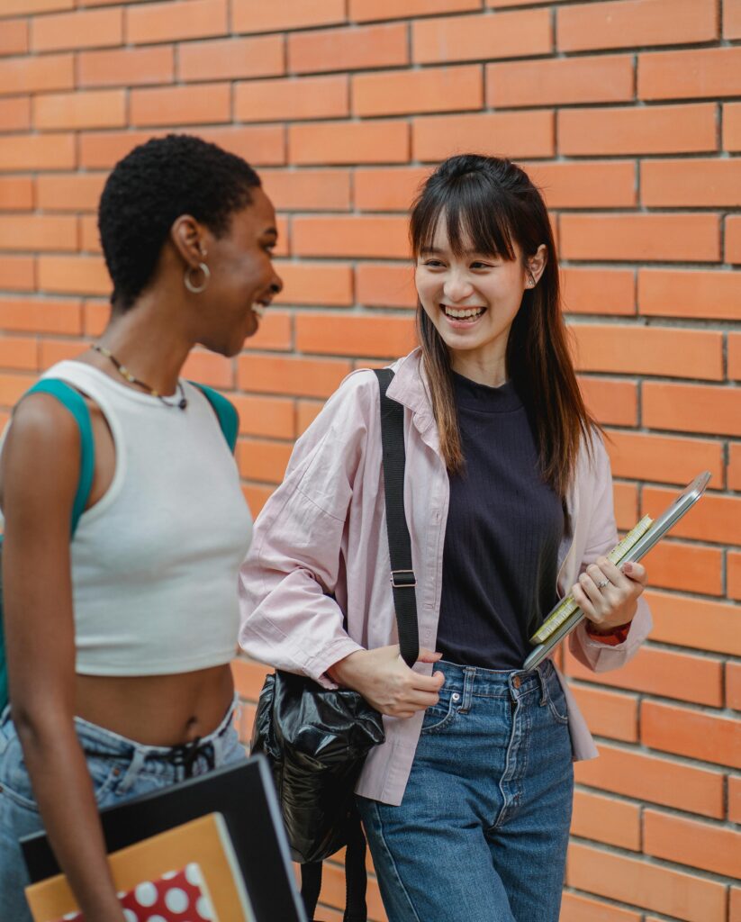 Laughing multiethnic female students wearing casual outfits strolling together against brick wall and chatting happily while looking at each other with smiles